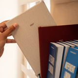A hand holds a beige 2027 edition of Papersmiths diary against a warm indoor background, with other blue and red volumes visible on a shelf.