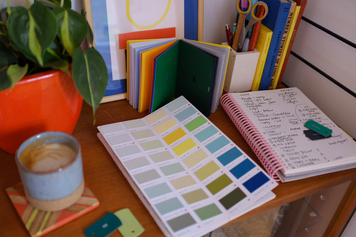 Color swatch book, coffee cup, and stationery on a desk with a plant and books in the background.
