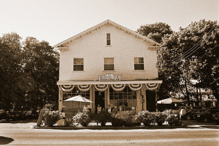 A sepia-toned photograph of a historic white wooden building with a storefront, bunting, outdoor umbrellas, trees, and parked cars along the street.