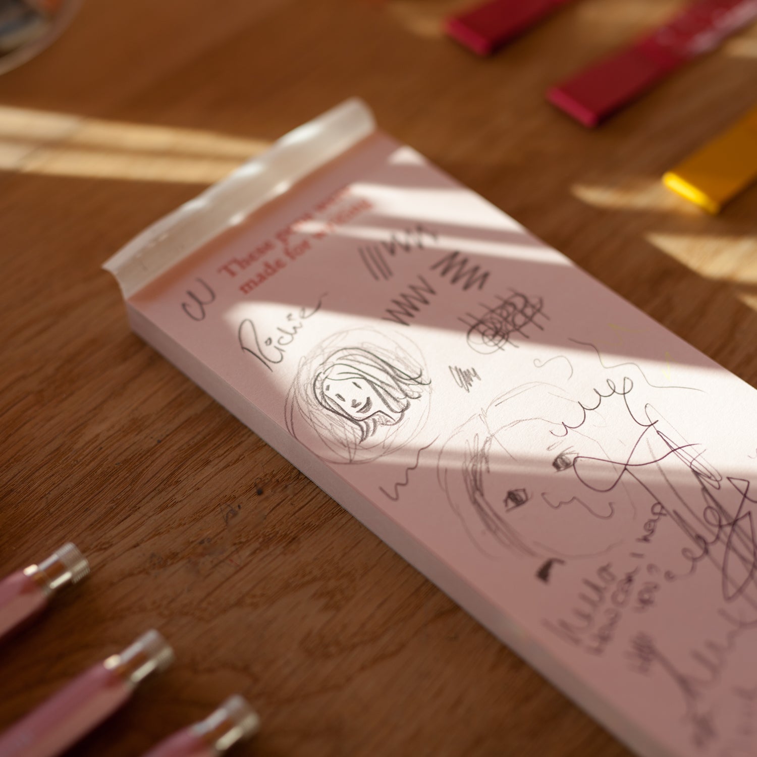 Pink notebook with doodles on a wooden surface, with pink pens nearby.