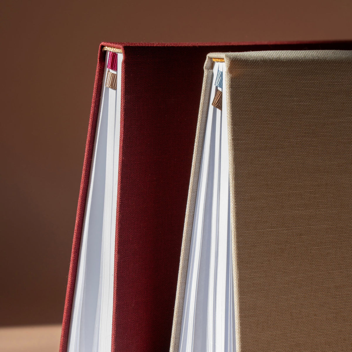 Close-up of a hardcover notebook or journal with a deep burgundy red fabric cover and white pages, standing next to another beige textured cover against a warm brown background.