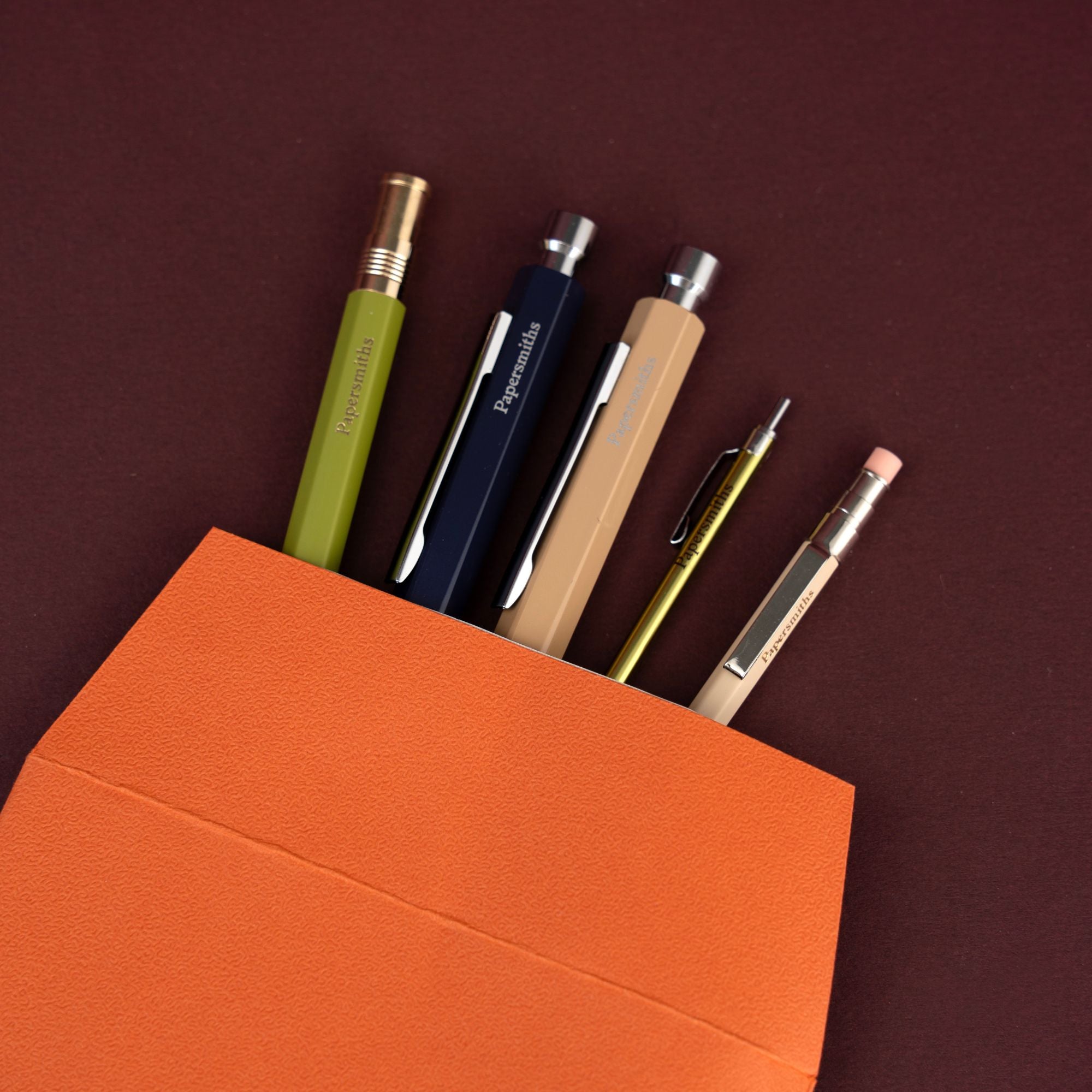 Five mechanical pencils in assorted colors (olive green, navy blue, beige, gold, and cream) arranged in an open bright orange envelope against a dark maroon background.