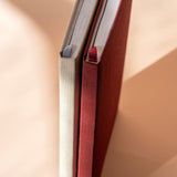 Two hardcover books or notebooks standing upright close together, showing their textured fabric spines in cream and deep red against a warm beige background.