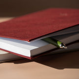 A close-up of a dark red fabric-covered notebook or journal with white pages, partially covered by a matching red cover, with a green pen resting across the edge on a warm beige surface.
