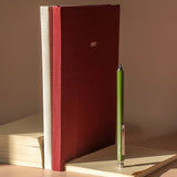 A dark red fabric-covered 2027 planner or journal stands upright on a beige surface, next to a green pen resting on a light notebook. The scene is lit by warm directional sunlight, casting soft shadows against a neutral beige background.