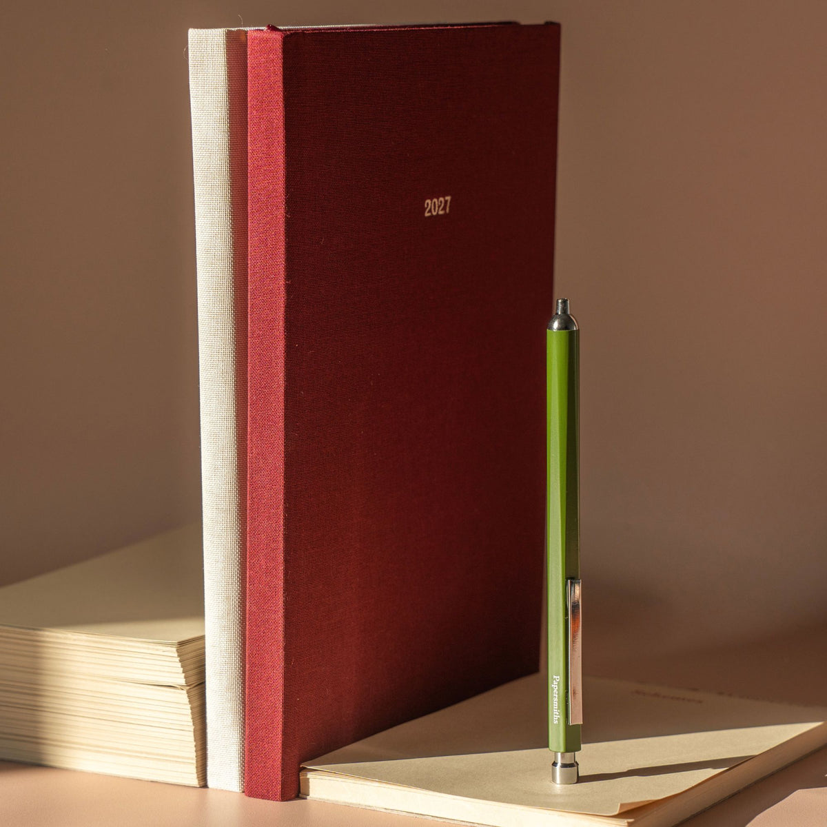 A dark red fabric-covered 2027 planner or journal stands upright on a beige surface, next to a green pen resting on a light notebook. The scene is lit by warm directional sunlight, casting soft shadows against a neutral beige background.