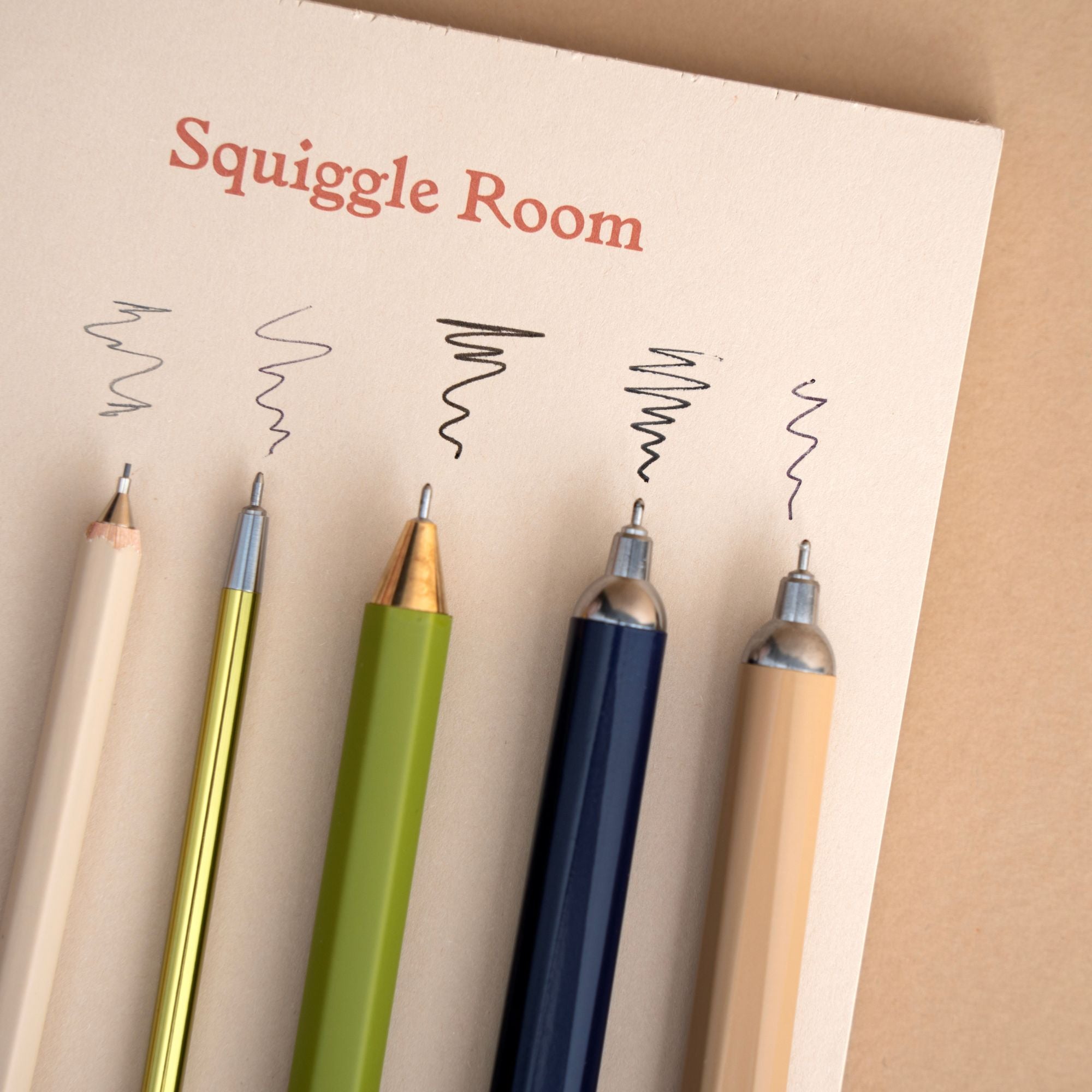 Four pens in beige, gold, green, and navy blue aligned on a cream-colored card that reads 'Squiggle Room,' with black squiggle marks above each pen.