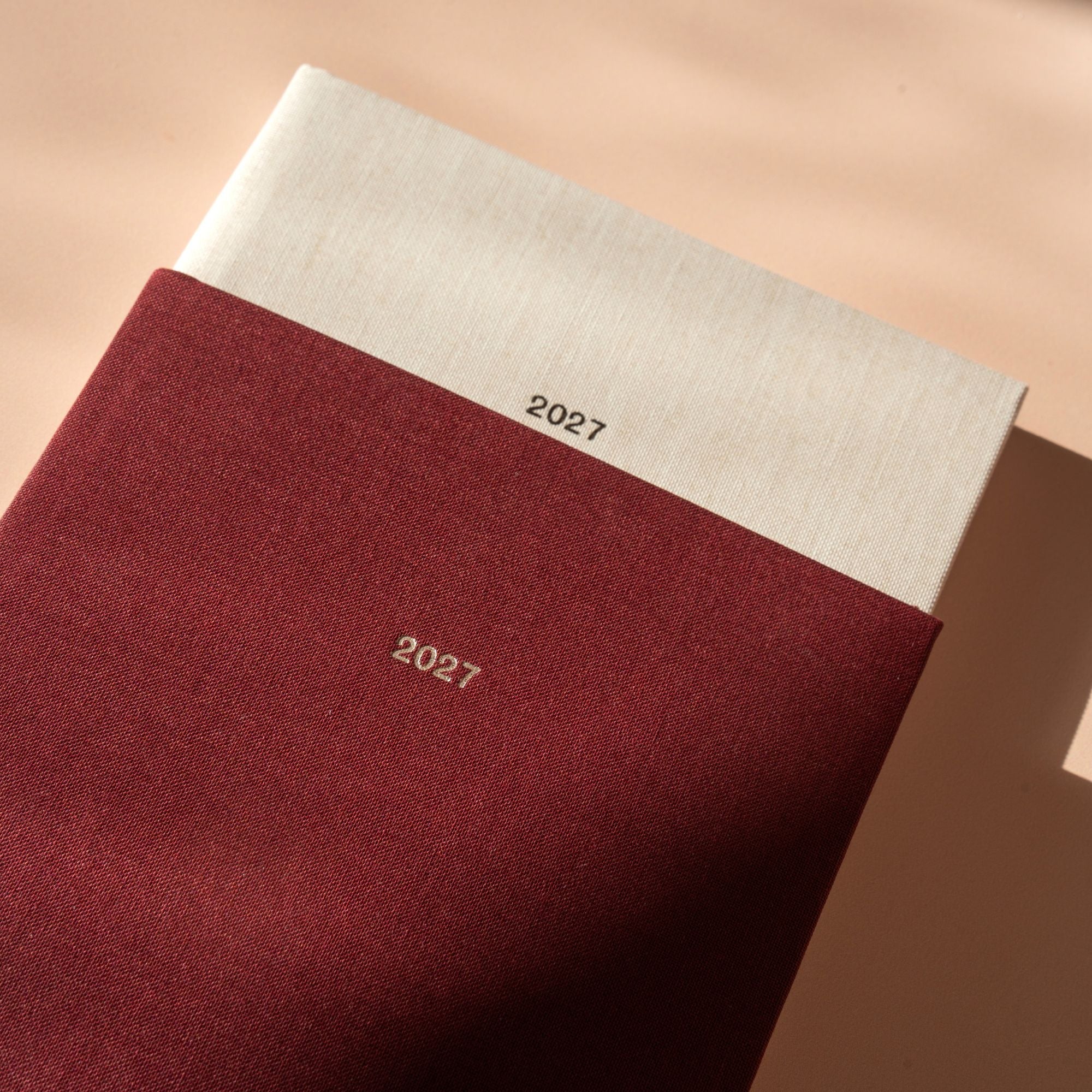 A close-up of a 2027 fabric-covered journal or planner, showing a deep burgundy cover with a white top flap and the year '2027' printed in gold. The photo is styled with warm, directional sunlight and soft shadows on a beige surface.
