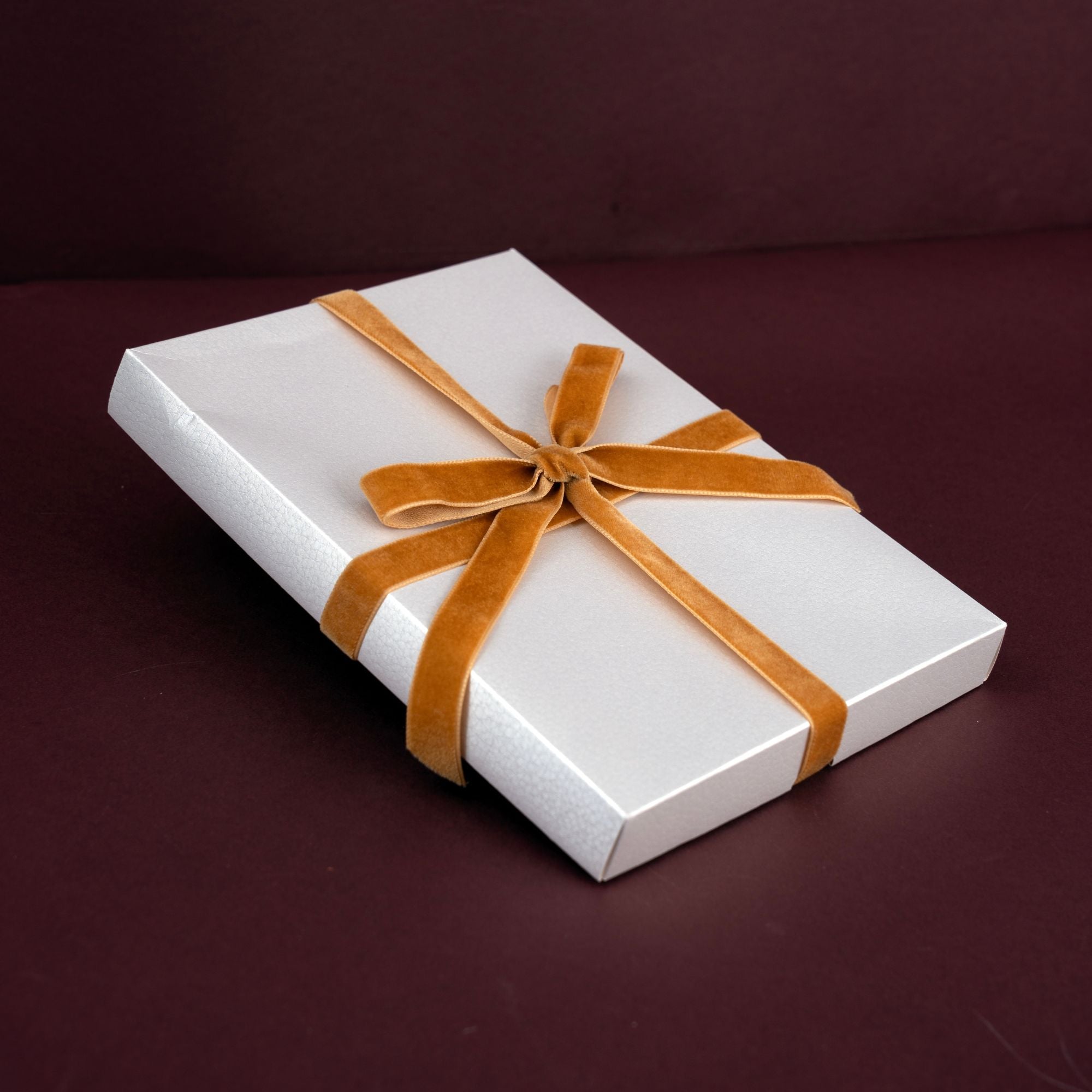 A square white gift box wrapped with a wide tan velvet ribbon tied in a bow, set against a dark burgundy background.