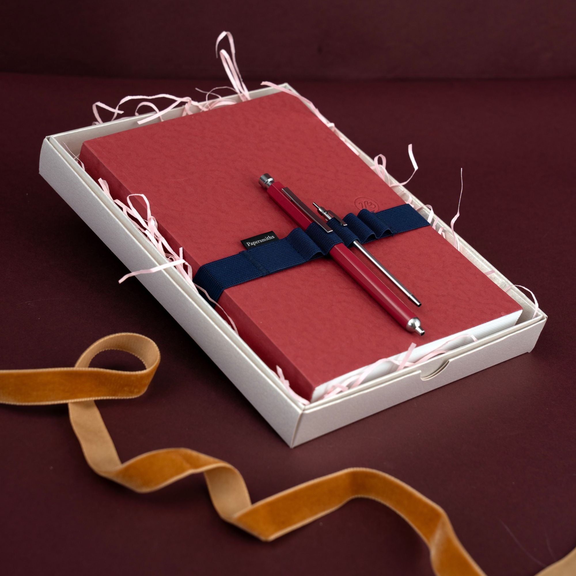 A red notebook with a navy blue elastic band and a matching red pen in a white gift box with shredded paper, set against a dark burgundy background with a gold ribbon in the foreground.