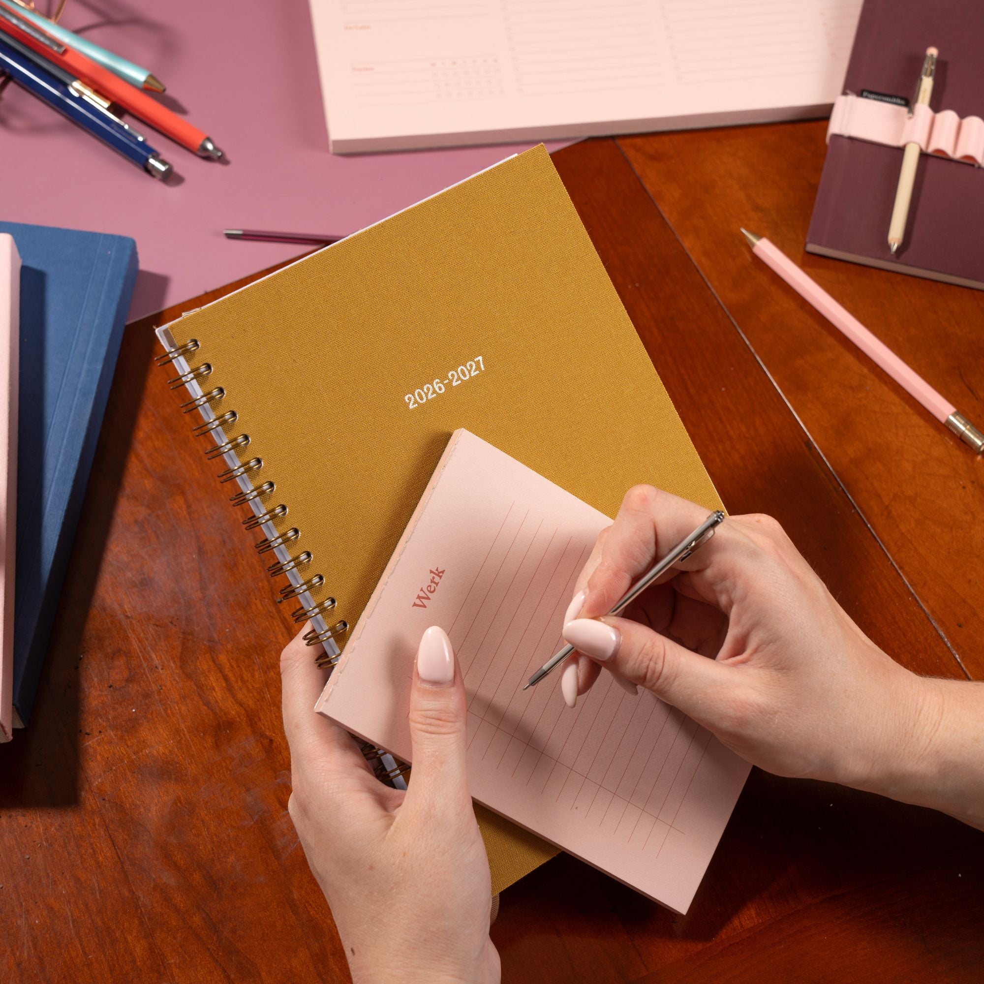 Person writing in a pink notebook with a 2026-2027 planner on a wooden table.